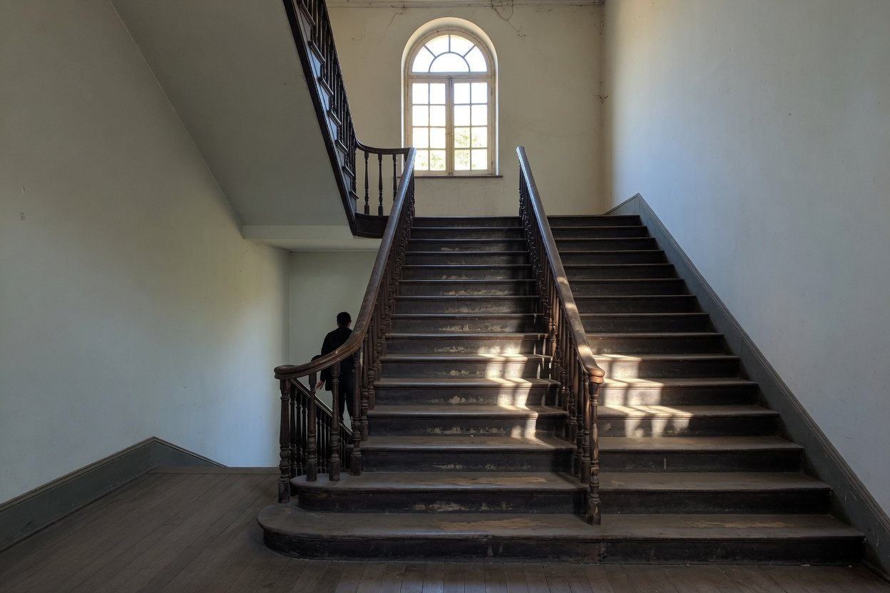 Escadaria de madeira em lances e patamares num predio publico antigo, subindo em direcao a uma janela clara
