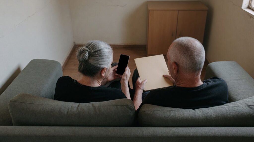 Casal de idosos no sofa escutando uma ligacao no viva-voz, com uma pasta fechada no colo
