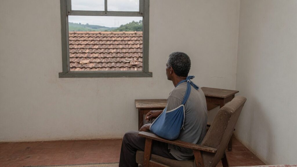 Homem com o braco na tipoia sentado numa poltrona junto a janela do quarto