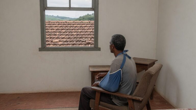 Homem com o braco na tipoia sentado numa poltrona junto a janela do quarto
